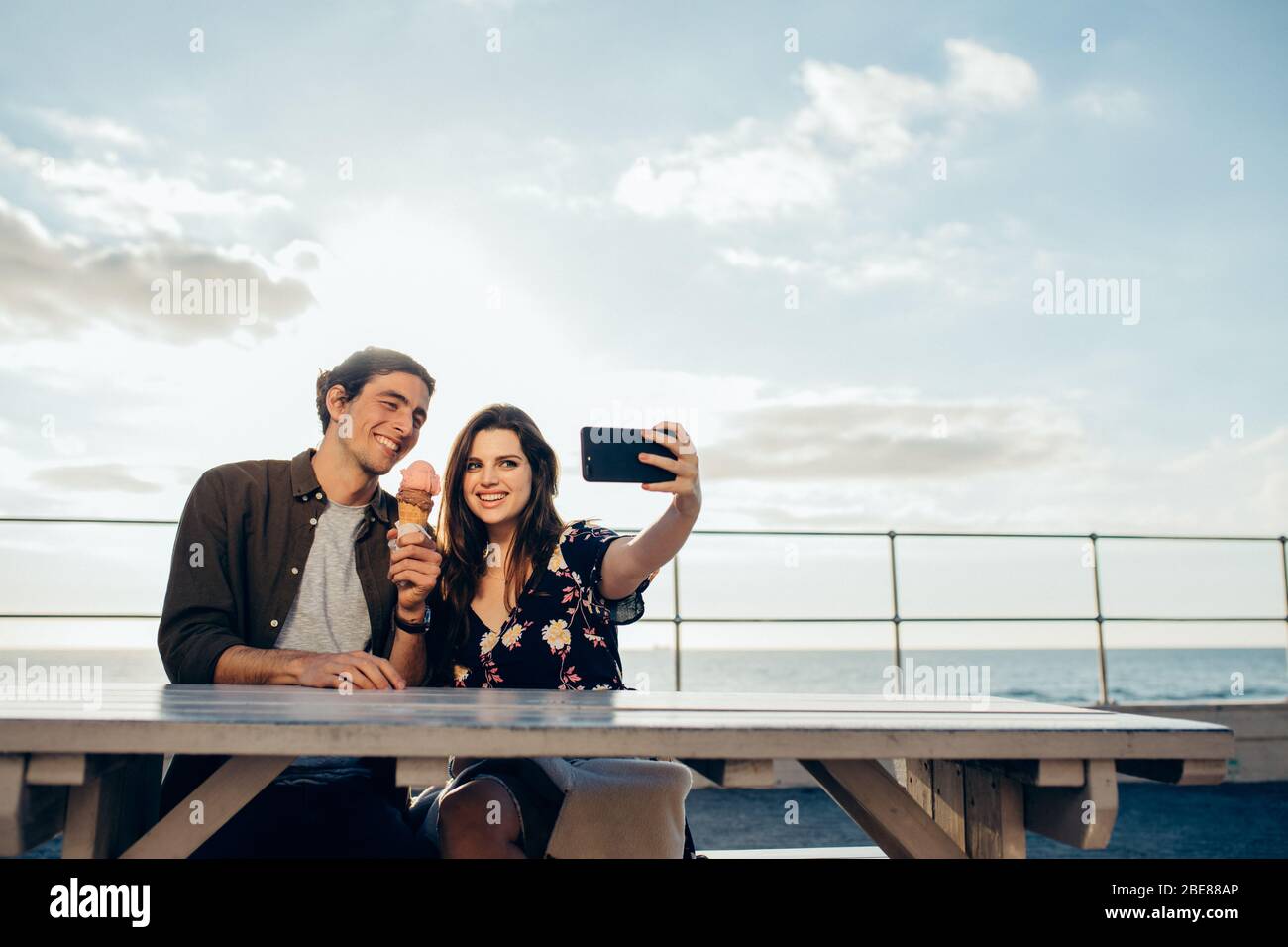 Femme prenant selfie avec smartphone avec son petit ami tenant une glace assis à l'extérieur. Couple prenant selfie à leur date. Banque D'Images
