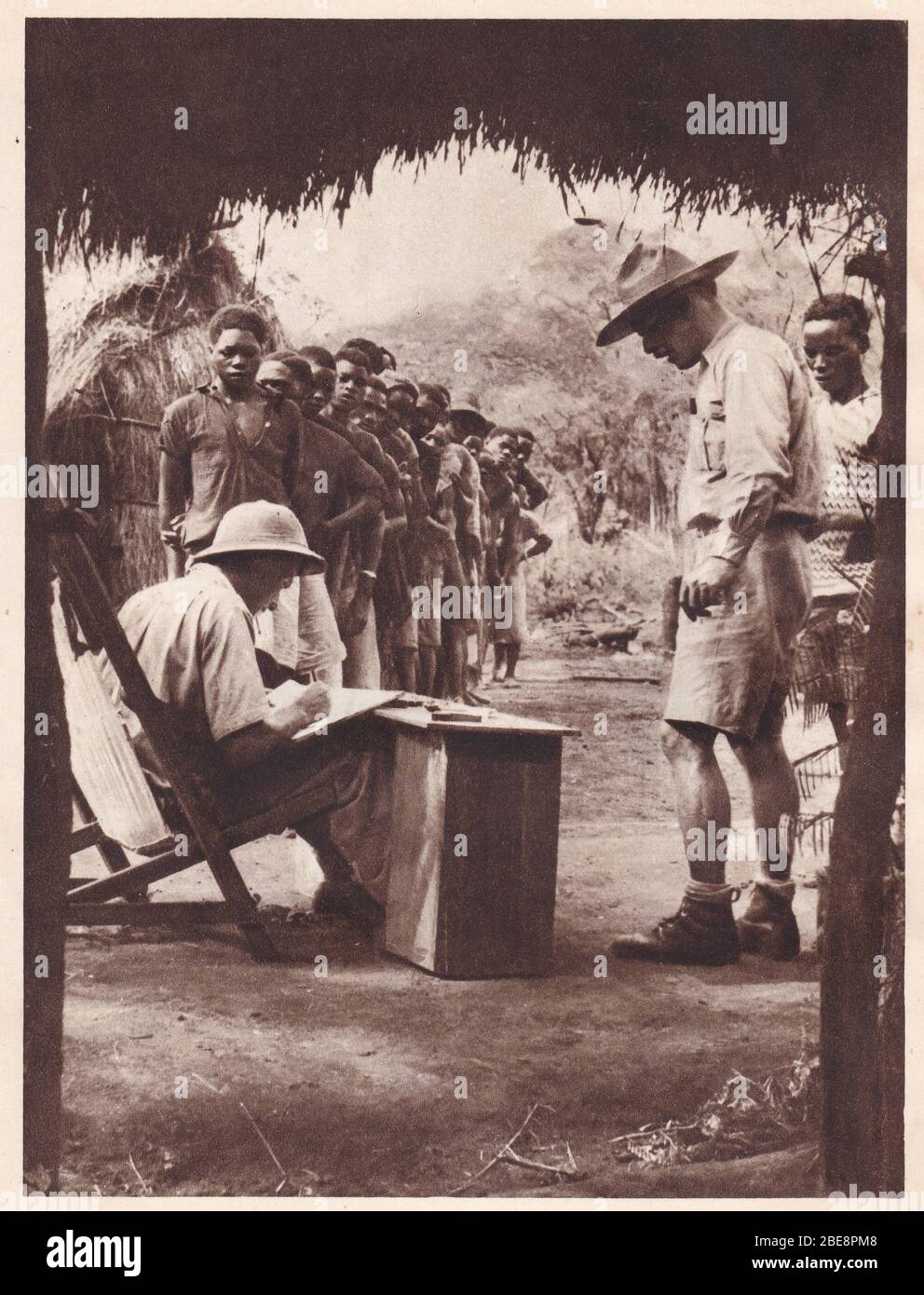 Photo noire et blanche vintage des travailleurs autochtones africains des années 1930, doublée pour recevoir leur salaire des hommes blancs en tenues de safari. Banque D'Images