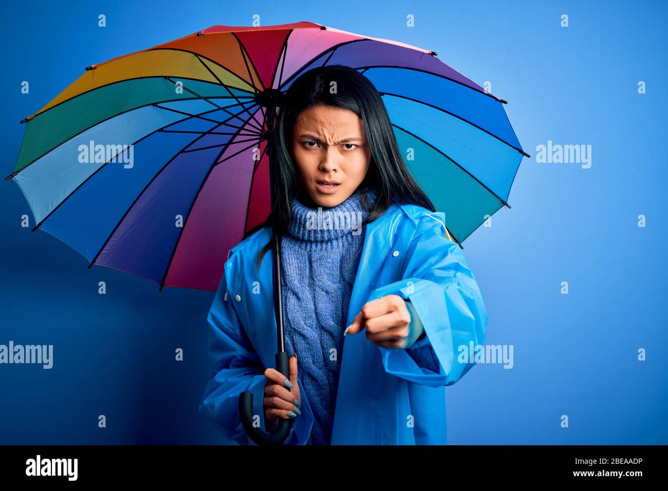 Une jeune femme chinoise portant un manteau de pluie portant un parapluie  sur un fond bleu isolé s'est énervé et frustrée de crier avec colère, esprit  fou et hurlant Photo Stock -