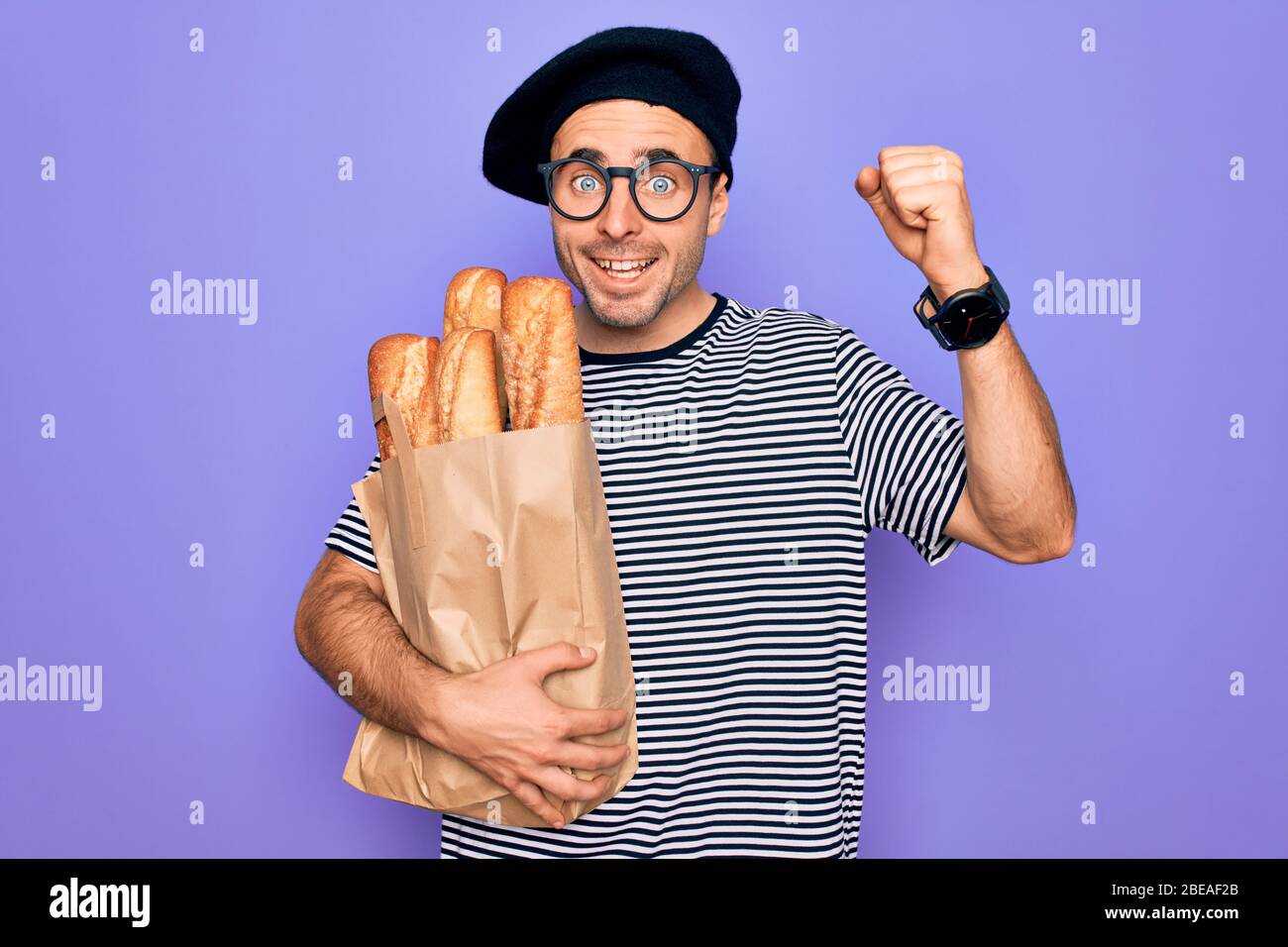 Jeune homme boulanger aux yeux bleus portant un sac de maintien en béret français avec du pain criant fier et célébrant la victoire et le succès très excité, Banque D'Images