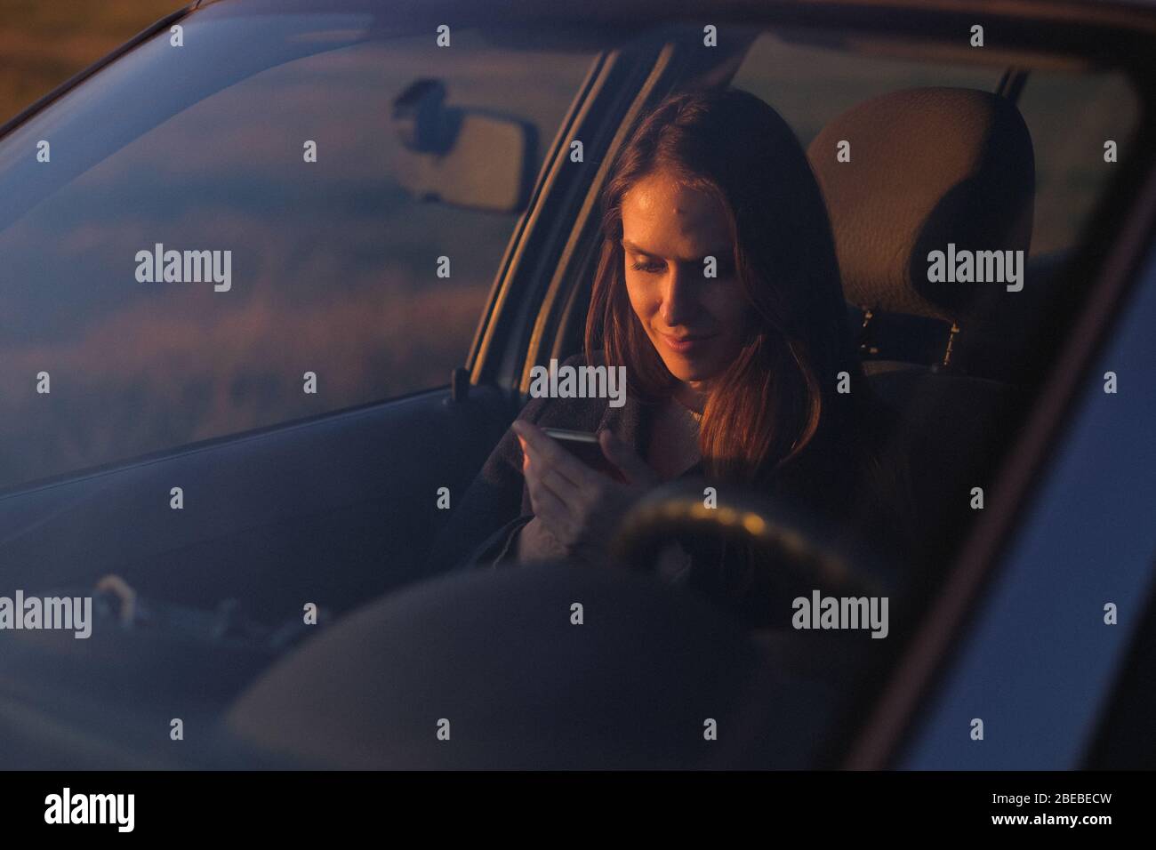 Une femme assise en voiture utilise un smartphone le soir avant le coucher du soleil. Banque D'Images
