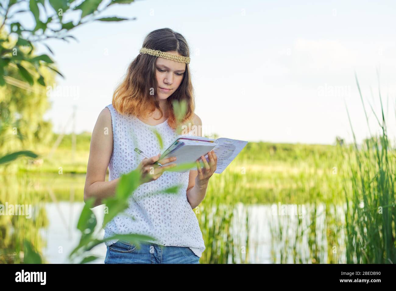 Belle fille romantique adolescent avec carnet personnel. Journal d'écriture et de lecture des femmes, arrière-plan nature pittoresque du lac surcultivé avec des plantes Banque D'Images