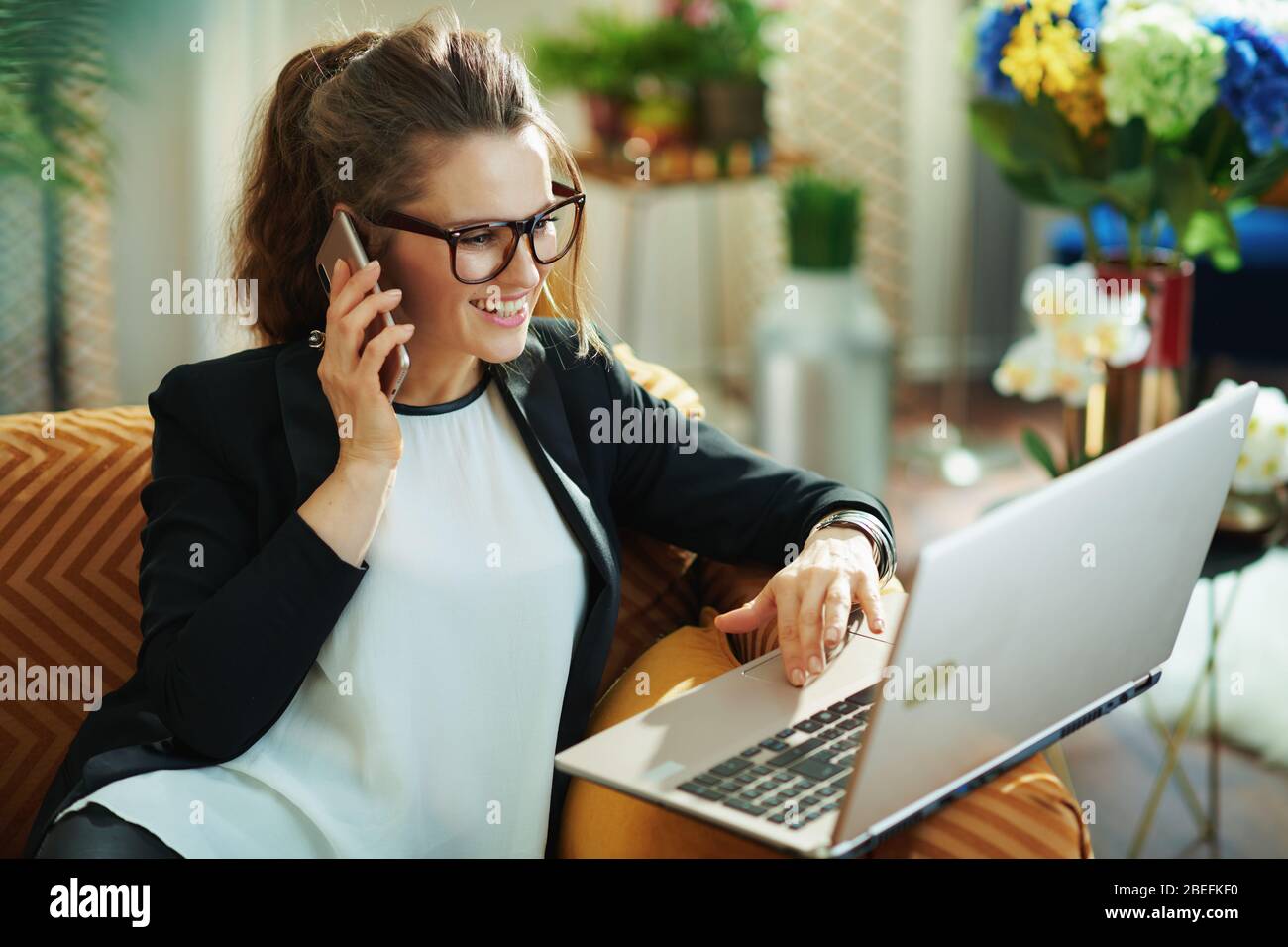 femme souriante et élégante avec chemisier blanc et veste noire avec ordinateur portable utilisant un téléphone portable à la maison moderne en journée ensoleillée. pavé tactile défilant Banque D'Images