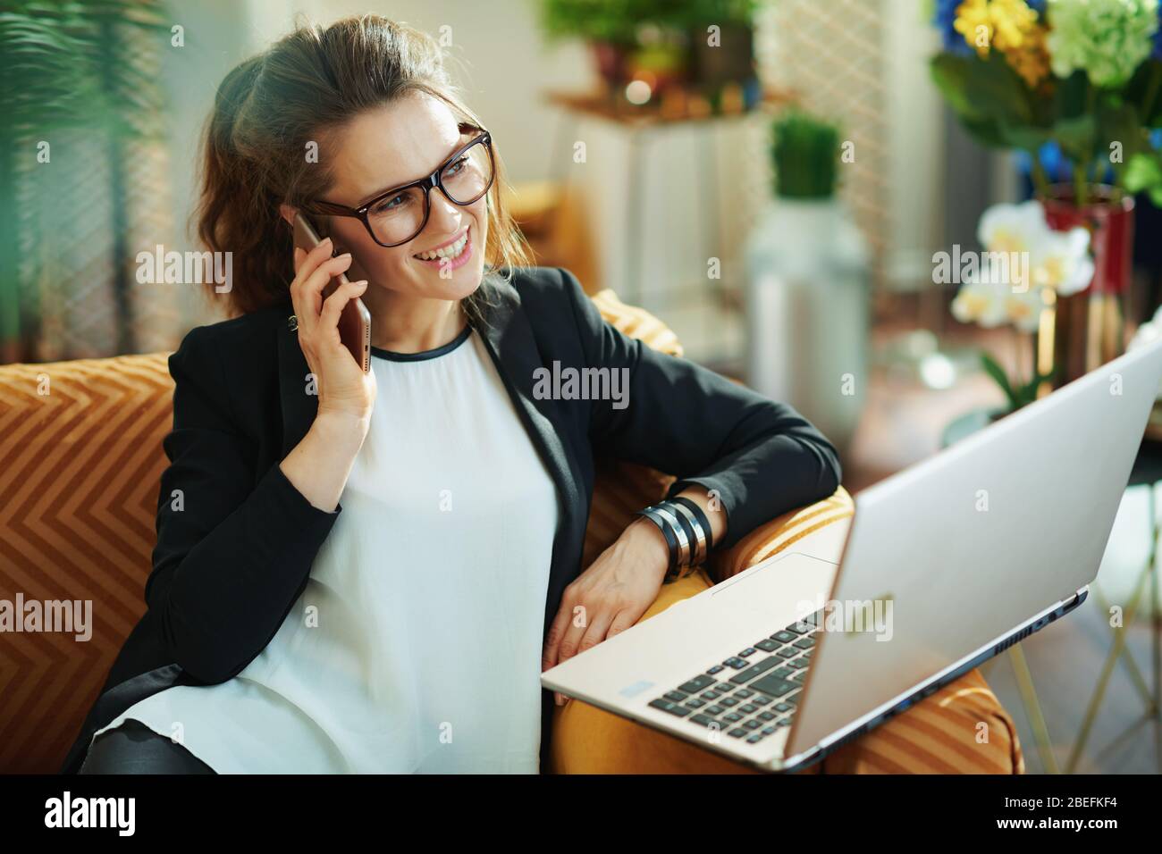 heureuse femme moderne avec chemisier blanc et veste noire avec ordinateur portable parlant sur un téléphone portable à la maison moderne en journée ensoleillée. Banque D'Images