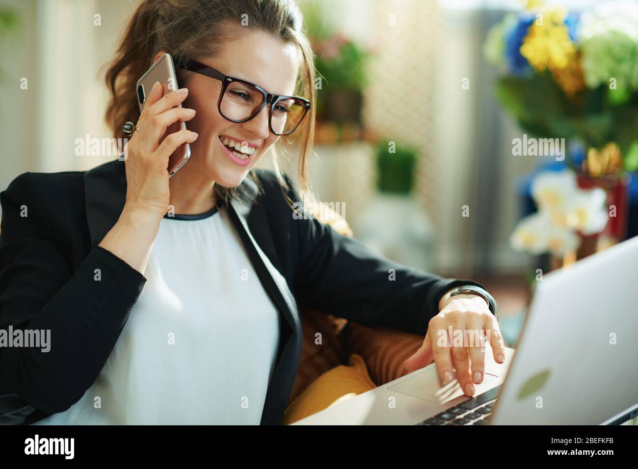 femme de ménage moderne souriante avec chemisier blanc et veste noire avec ordinateur portable utilisant un téléphone mobile dans le salon moderne en journée ensoleillée. saisie au clavier Banque D'Images