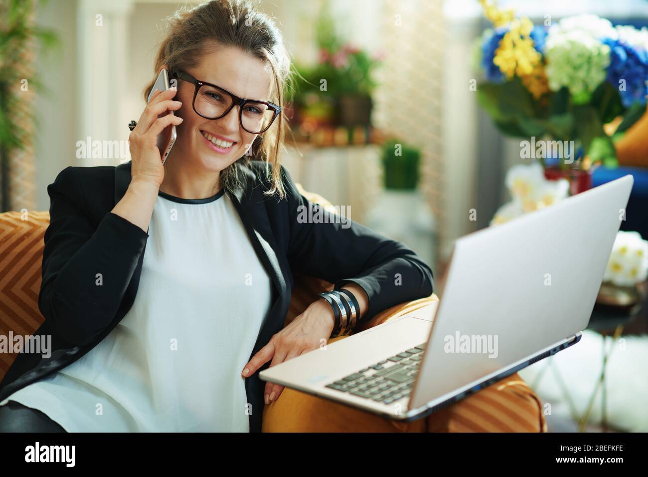 bonne femme de ménage de 40 ans avec chemisier blanc et veste noire avec ordinateur portable parlant sur un téléphone mobile dans la maison moderne en journée ensoleillée. Banque D'Images