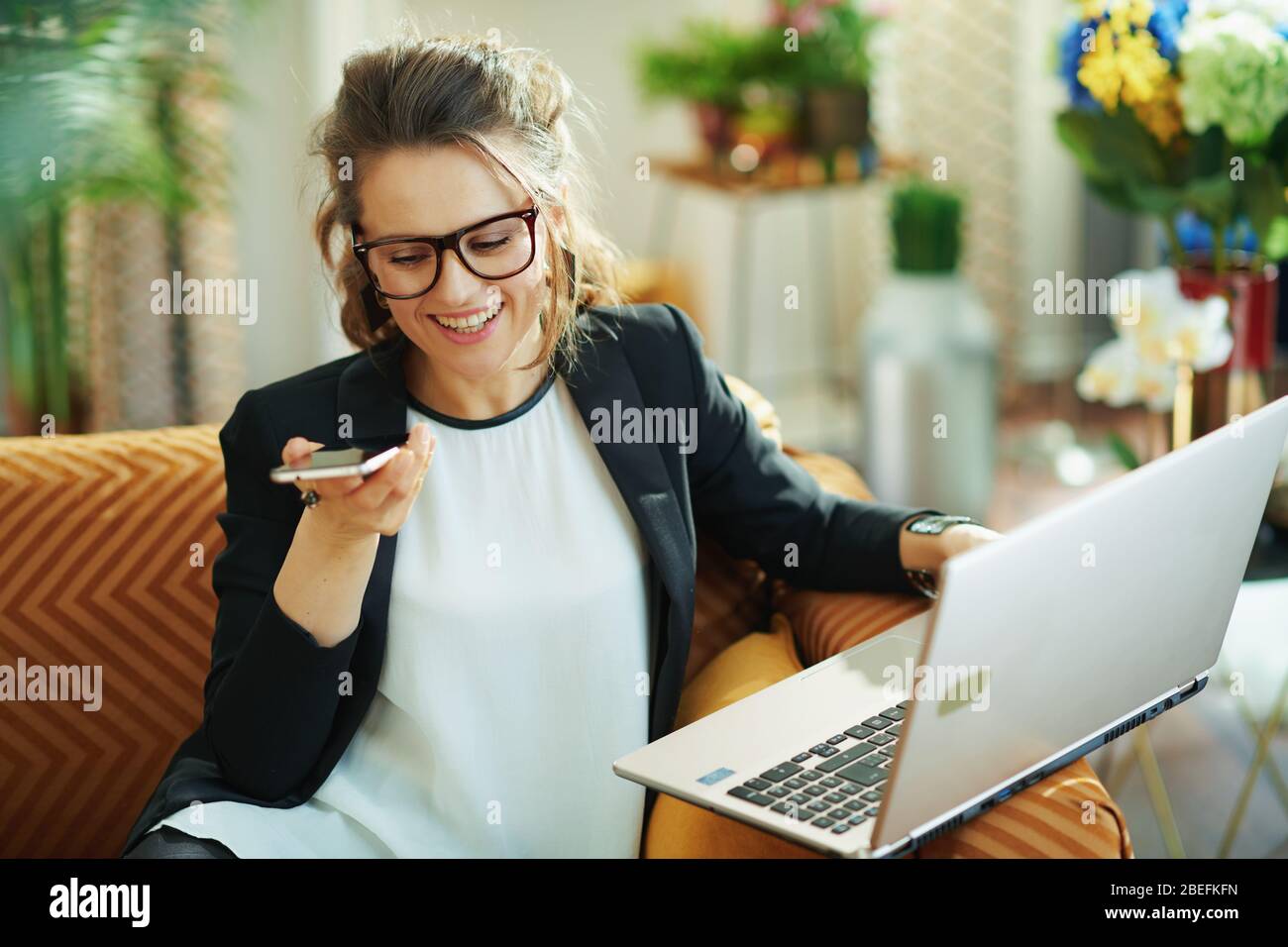 femme de ménage souriante et élégante de 40 ans avec chemisier blanc et veste noire avec ordinateur portable parlant sur un téléphone mobile dans la maison moderne en journée ensoleillée. Banque D'Images