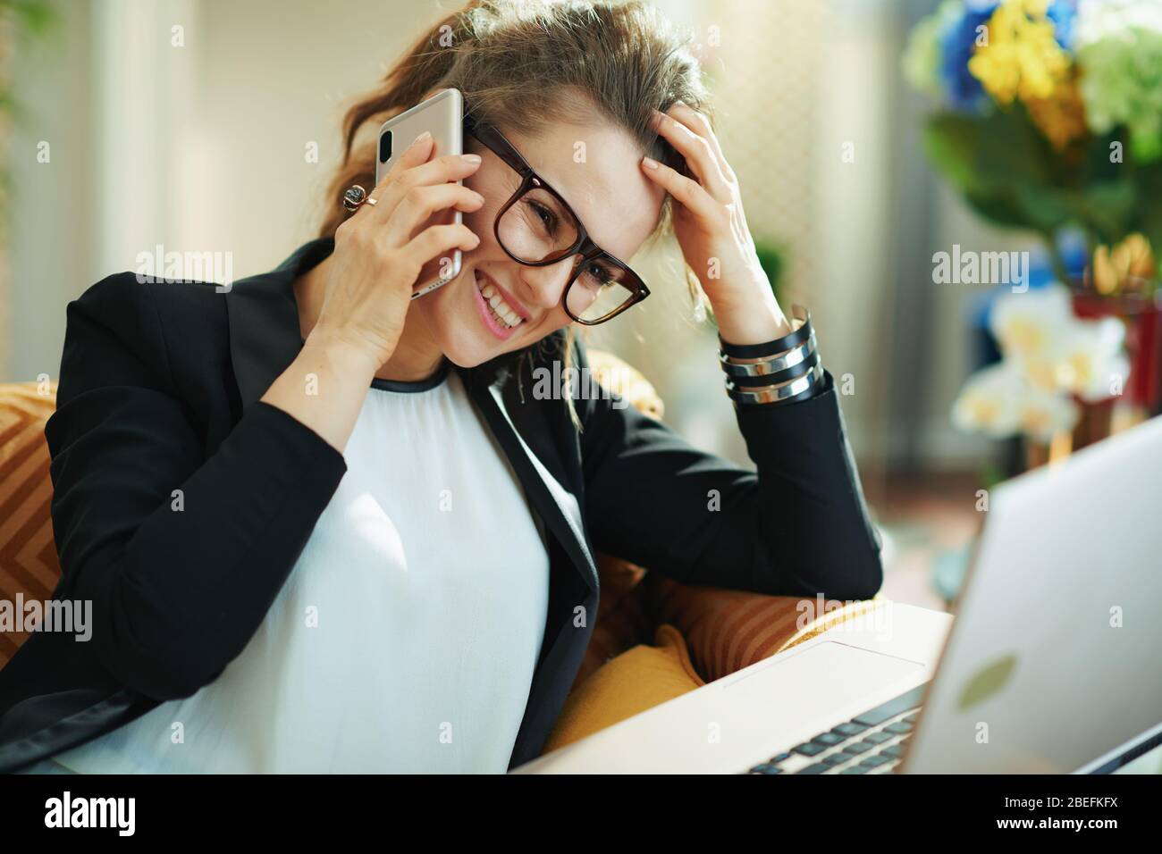 heureuse jeune femme en chemisier blanc et veste noire avec ordinateur portable parlant sur un téléphone portable à la maison moderne en journée ensoleillée. Banque D'Images
