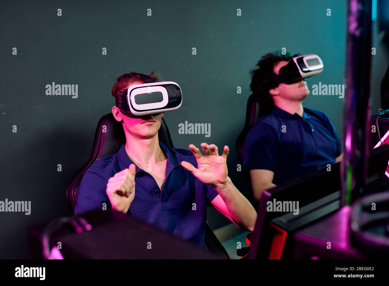 Curieux jeunes hommes dans les mêmes tee-shirts assis dans le sombre e-sport club et utilisant des simulateurs de réalité virtuelle tout en jouant jeu vidéo réseau Banque D'Images