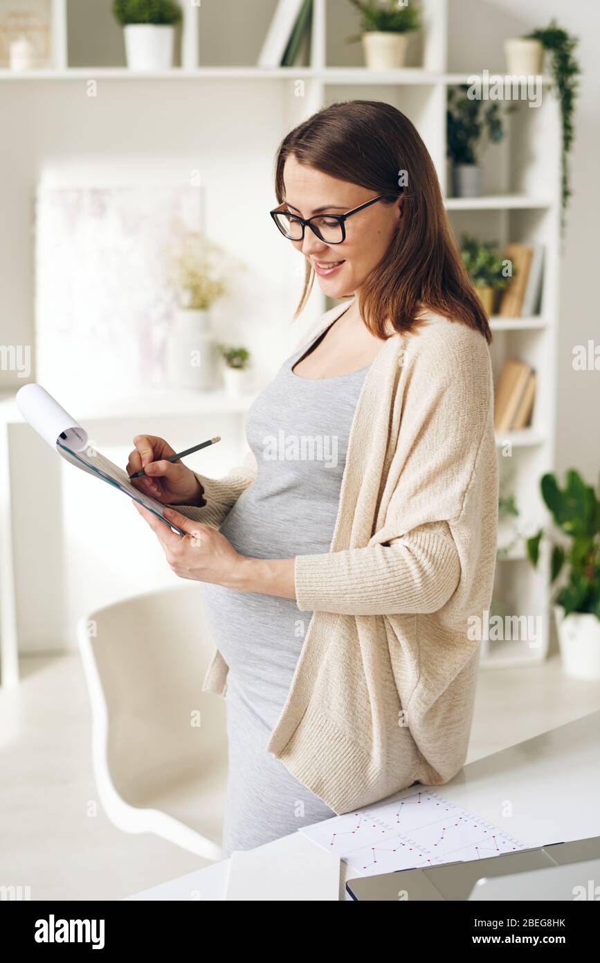 Femme enceinte souriante et élégante dans un gilet et des lunettes se tenant au bureau et prenant des notes dans des papiers tout en l'analysant au bureau à domicile Banque D'Images