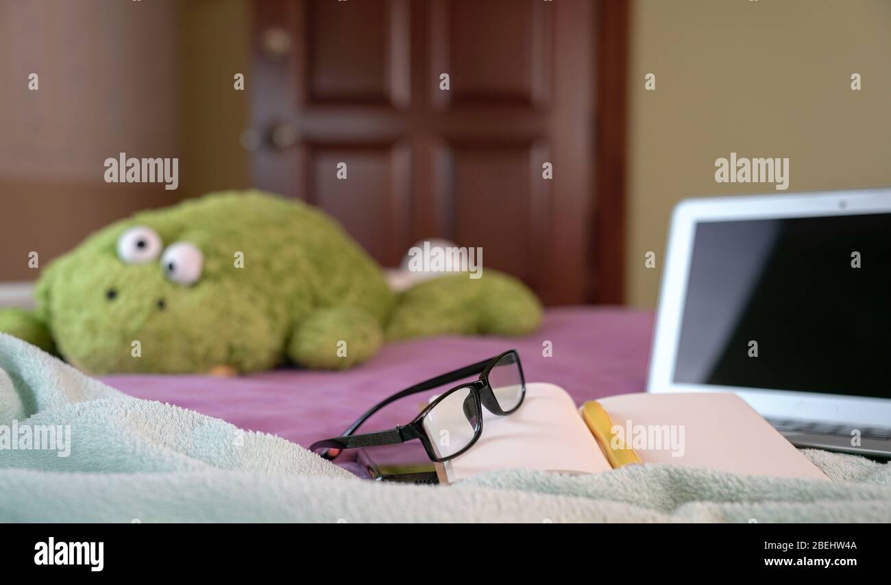 Ordinateur portable avec bloc-notes, stylo, lunettes de lecture et jouet doux sur le dessus du lit dans la chambre. Travailler à la maison. Se concentrer sur les lunettes. Banque D'Images