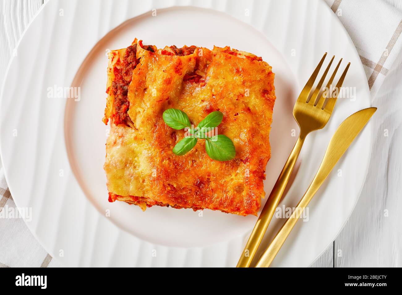 Cannelloni à la viande cuit avec sauce tomate et fromage mozzarella fondu et basilic frais sur une plaque blanche avec couverts sur fond en bois blanc, Banque D'Images