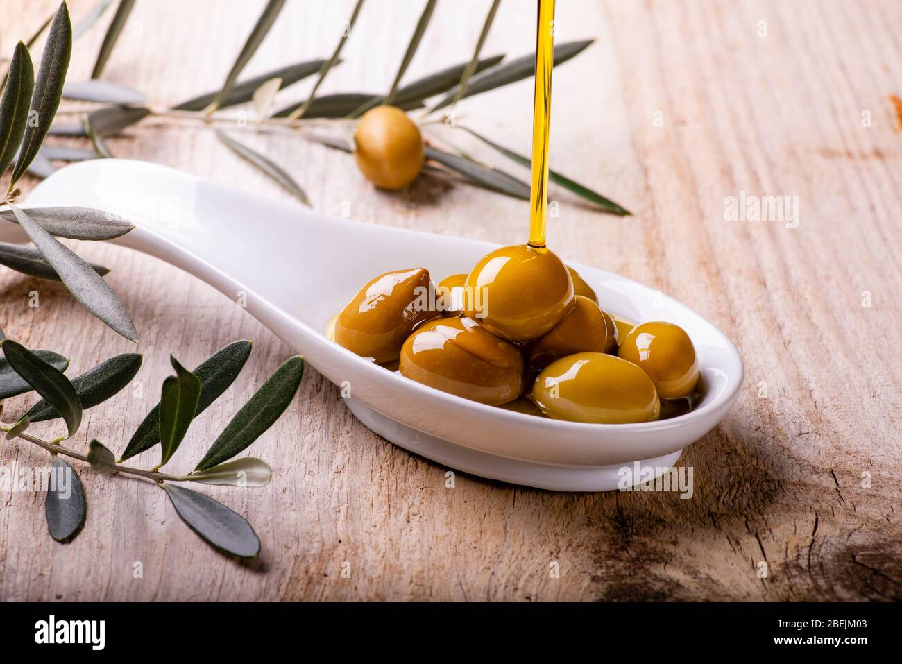 L'huile d'olive extra vierge s'écoule dans le bol en céramique rempli d'olives vertes sur la table en bois rustique Banque D'Images
