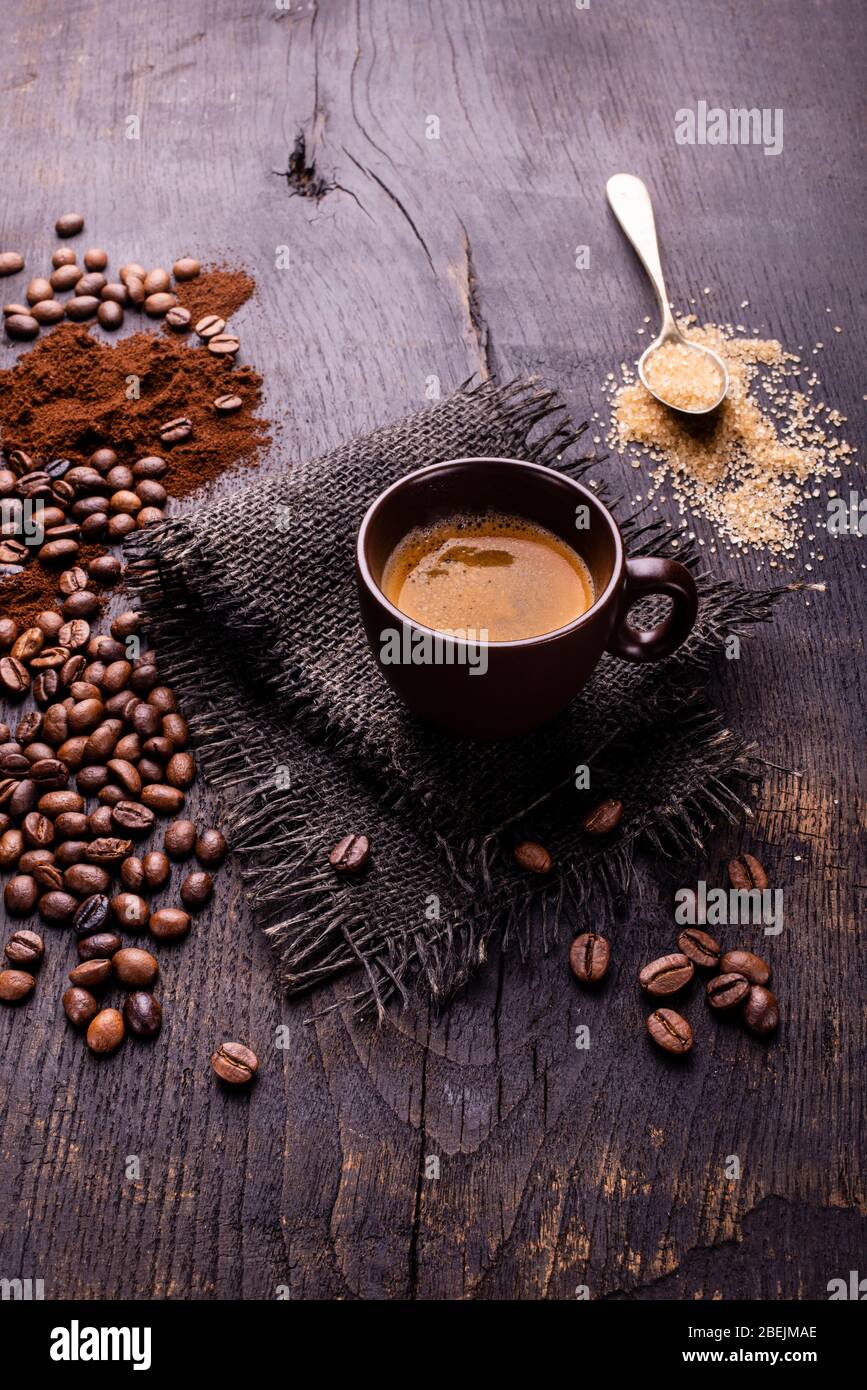 Sur la table en bois brut et sombre, une tasse de café avec une cuillerée de sucre brun et de grains de café torréfiés. Banque D'Images