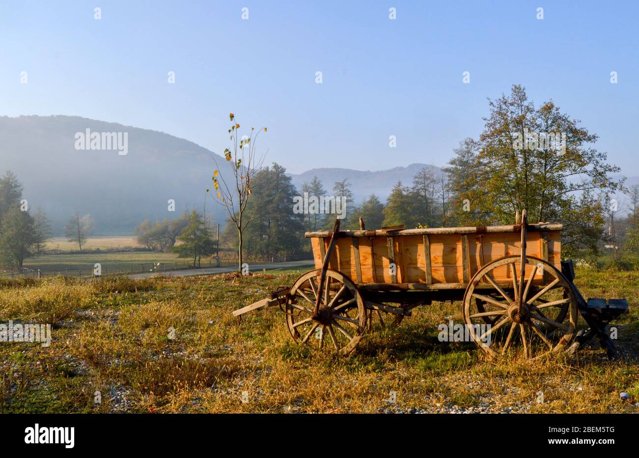 Paysage rural, quelque part dans les montagnes avec lumière chaude de lever de soleil sur un champ avec herbe et arbres, une vieille charrette en bois avec roues anciennes, rustique Banque D'Images