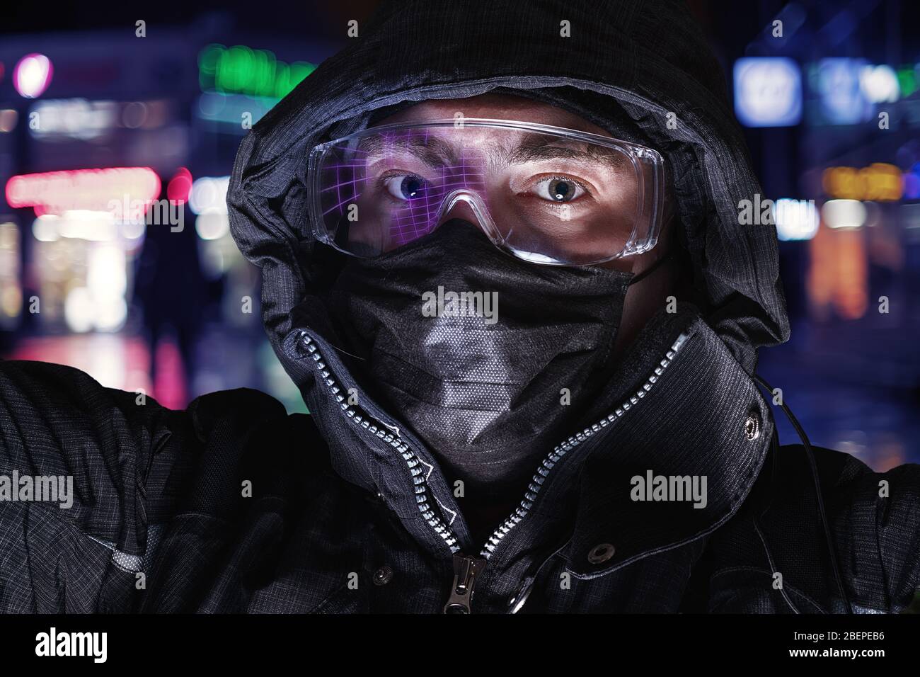 Homme sérieux dans les lunettes de sécurité, le masque médical et la capuche contre la rue éclairée. Portrait de type selfie Banque D'Images