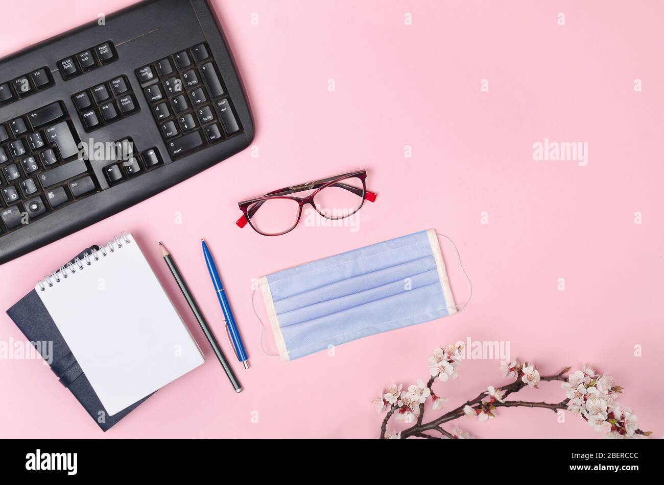 Branches fleuries avec un masque médical, un clavier, un bloc-notes et des lunettes sur un fond rose. Coronavirus COVID-19 Banque D'Images