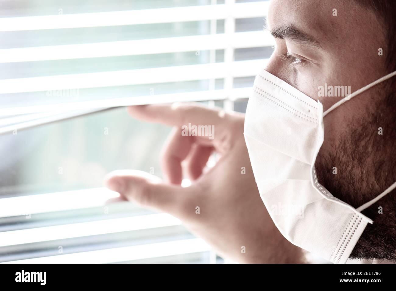 Photo d'un jeune homme blanc avec un masque médical regardant hors de la fenêtre avec jalousie pendant la quarantaine du coronavirus. Coronavirus, éclosion de COVID-19. Verrouillage, empêcher le concept de grippe. Banque D'Images
