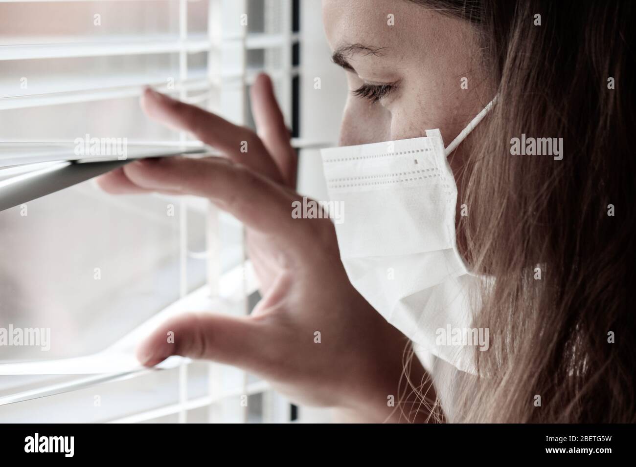 Photo d'une jeune femme blanche avec un masque médical regardant hors de la fenêtre avec jalousie pendant la quarantaine du coronavirus. Coronavirus, éclosion de COVID-19. Verrouillage, empêcher le concept de grippe. Banque D'Images