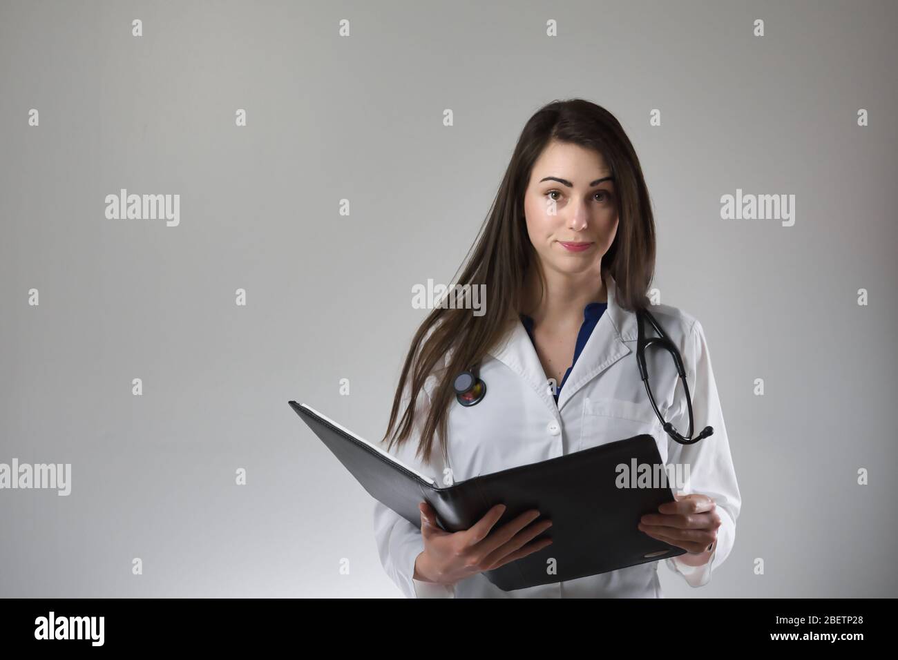 Professionnel de la santé féminin examinant le dossier patient isolé sur fond gris. Stéthoscope autour du cou, blouse blanche sur et boutonnée. Banque D'Images