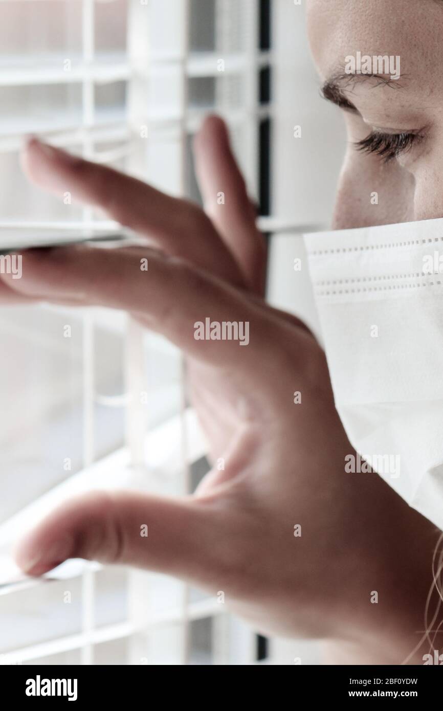 Photo d'une jeune femme blanche avec un masque médical regardant hors de la fenêtre avec jalousie pendant la quarantaine du coronavirus. Coronavirus, éclosion de COVID-19. Verrouillage, empêcher le concept de grippe. Banque D'Images