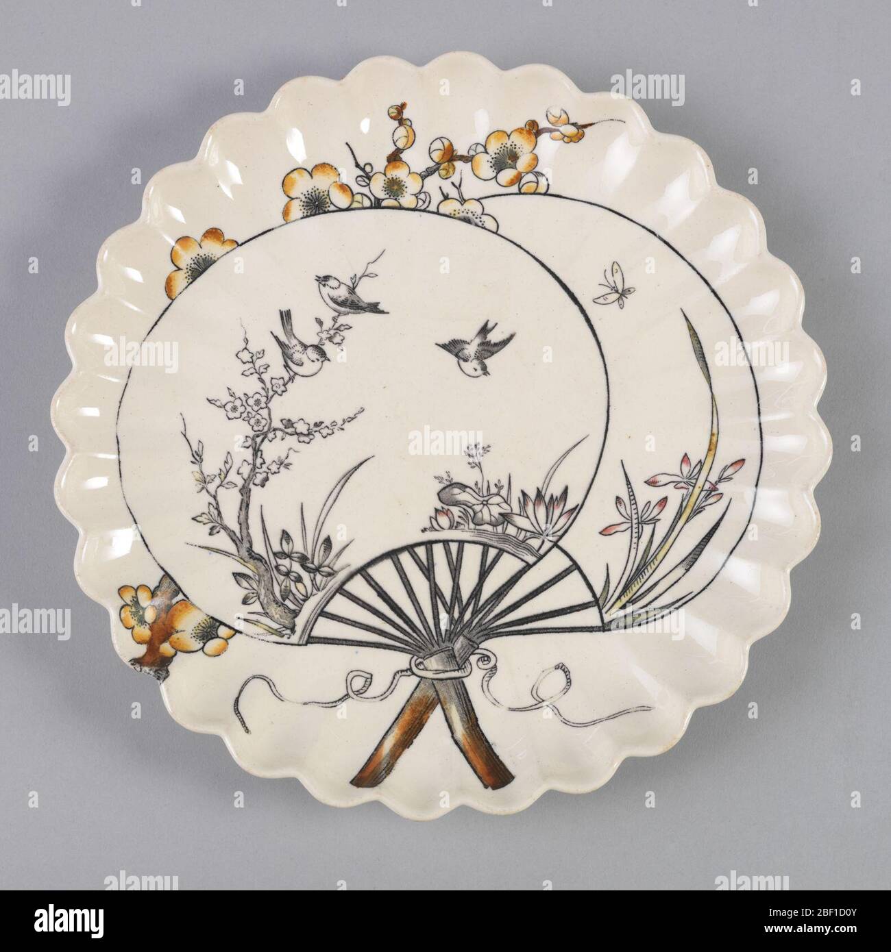 Plaque. Forme circulaire avec bord arrondi en hauteur, le sol crème avec décoration imprimée de transfert d'inspiration japonaise en noir et tons de brun et jaune montrant deux fans avec des scènes d'oiseaux et un papillon parmi les plantes et les branches fleuries; les fans super Banque D'Images