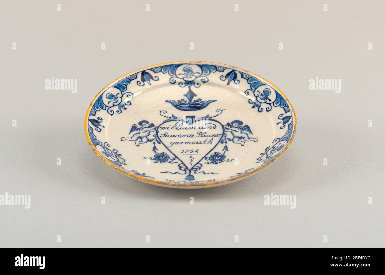 Plaque de mariage. Plaque circulaire avec rebord en haut; dôme en forme de dos, aplati au centre; peinte en bleu sous-glacis et jaune sur blanc avec un coeur surmonté d'une couronne et entouré de putti et de fleurs, inscrite à l'intérieur de 'William et / Suanna Plumer / Yarmouth / 1754'; Banque D'Images