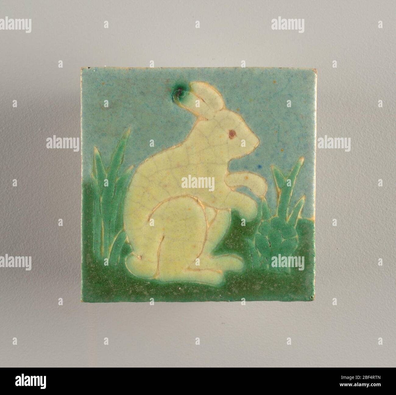 Mosaïque. Corps en faïence gris-blanc avec motif moulé sous pression de croûtage de lapin dans l'herbe. Bleu clair clair et vert foncé, et jaune pâle-vert mat glise définie par des cloisons surélevées. Banque D'Images