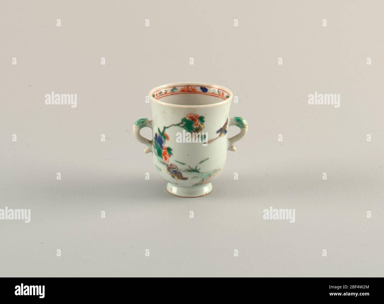 Bécher. Bécher à pied de famille verte avec deux poignées à forme de dauphin. Fleurs peintes avec canard; bande peinte d'ornement à l'intérieur du bord. Banque D'Images