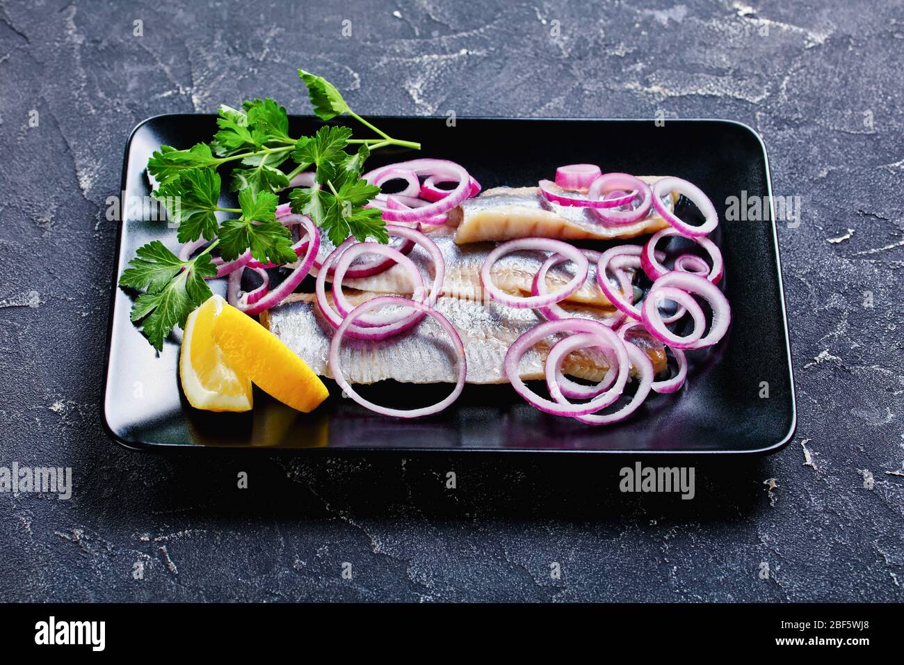 Cuisine scandinave hareng salé - poisson gras frais servi sur une plaque noire avec rondelles d'oignon rouge, persil et quartiers de citron avec couverts sur un c foncé Banque D'Images