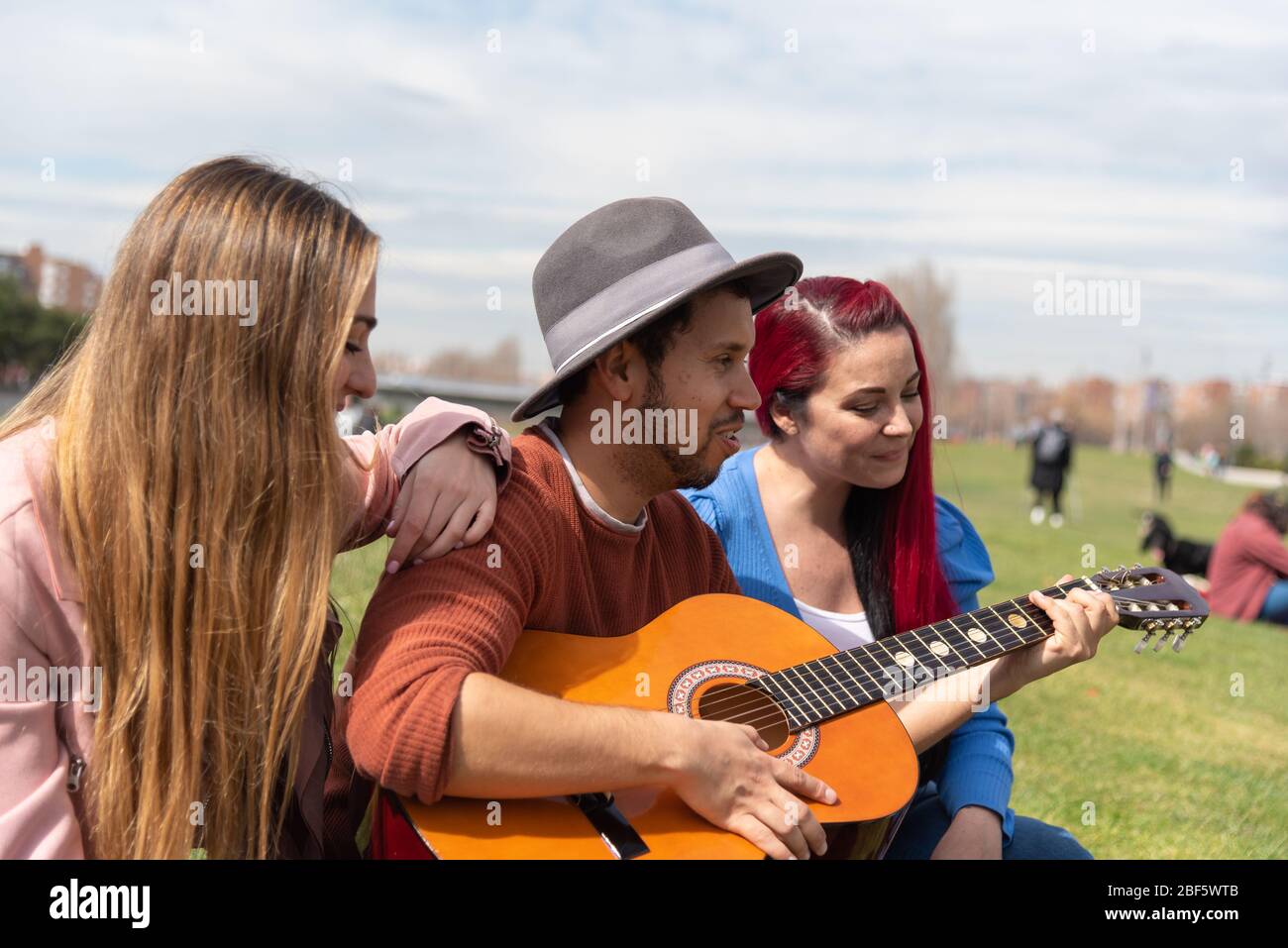 Un garçon hispanique dans un chapeau joue de la guitare avec deux filles caucasiennes dans un parc urbain Banque D'Images