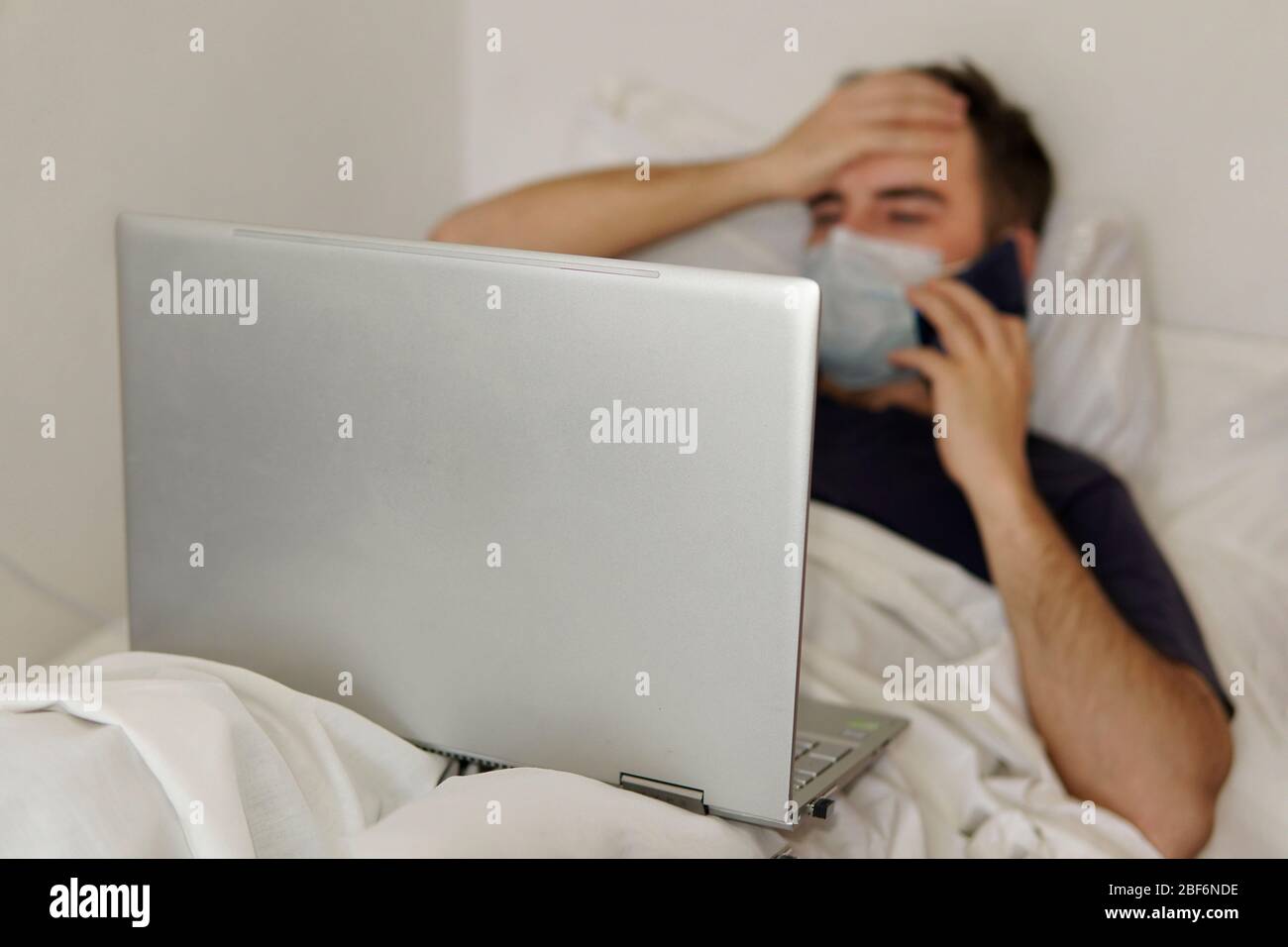 Un homme adulte est allongé dans un lit d'hôpital avec un ordinateur portable tenant sa tête. Concept de travail à distance pendant la pandémie du coronavirus Banque D'Images