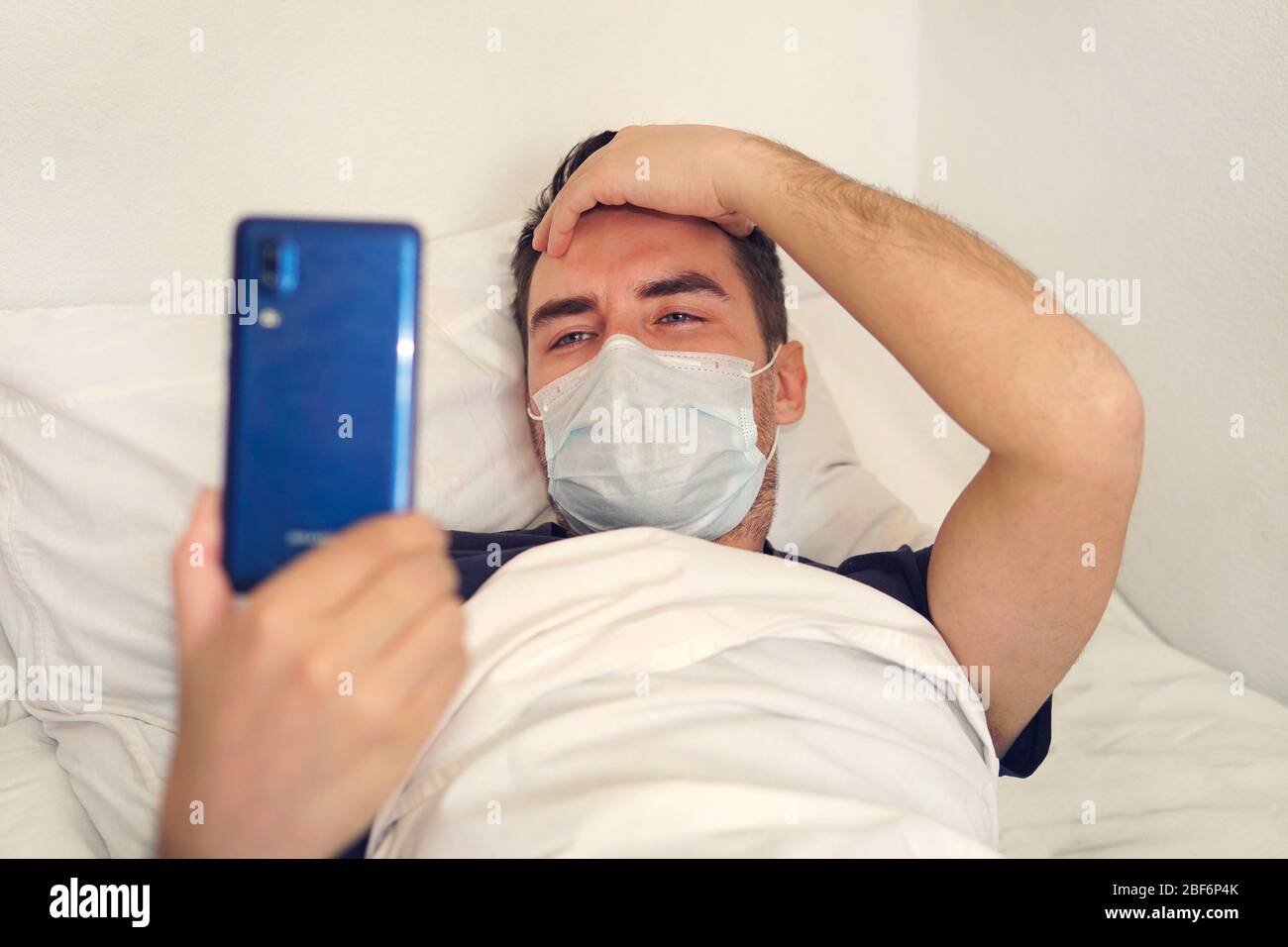 Temps d'ennui en quarantaine. Un jeune homme est traité dans une chambre d'hôpital. Le patient regarde l'écran du smartphone tout en étant allongé sur un lit blanc Banque D'Images