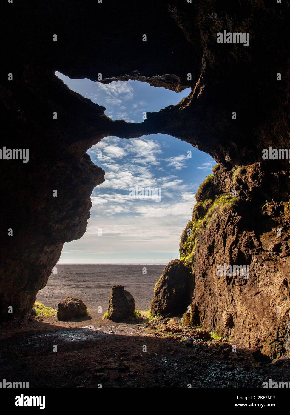 L'intérieur d'une grotte de Hjörleifshöfði, un inselberg en Islande, donne sur une plaine ensoleillée de sable volcanique, avec un ciel bleu et des nuages blancs Banque D'Images