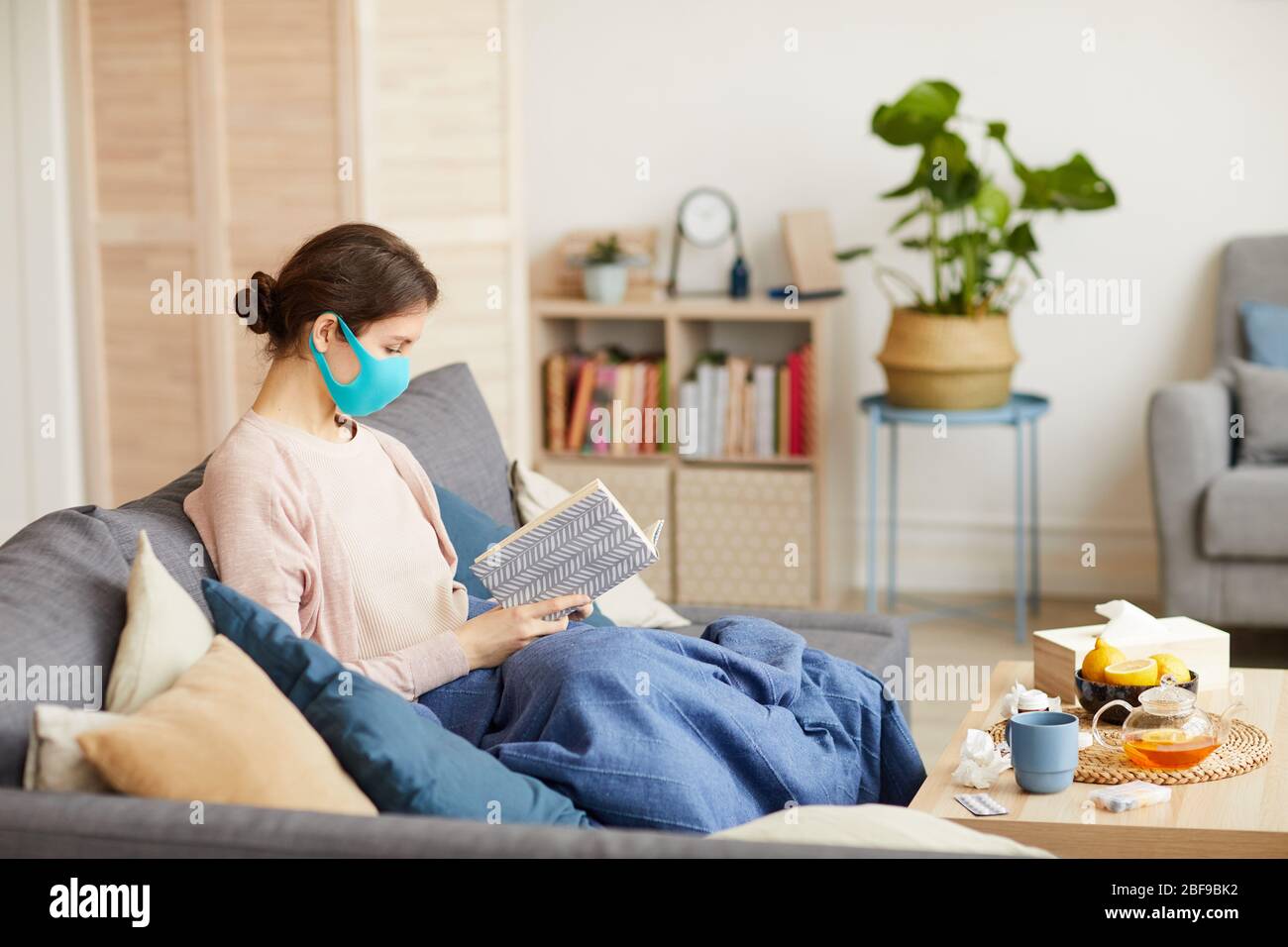 Malade femme portant un masque de protection assis sur un canapé avec livre elle est en congé de maladie et assis à la maison Banque D'Images