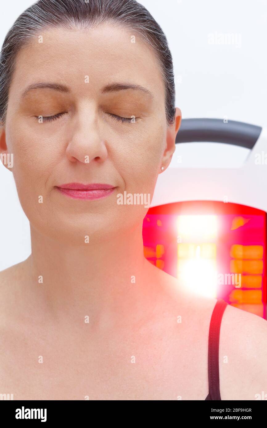 Femme souriante avec fibromyalgie devant la lampe infrarouge, traitement thermique. Banque D'Images