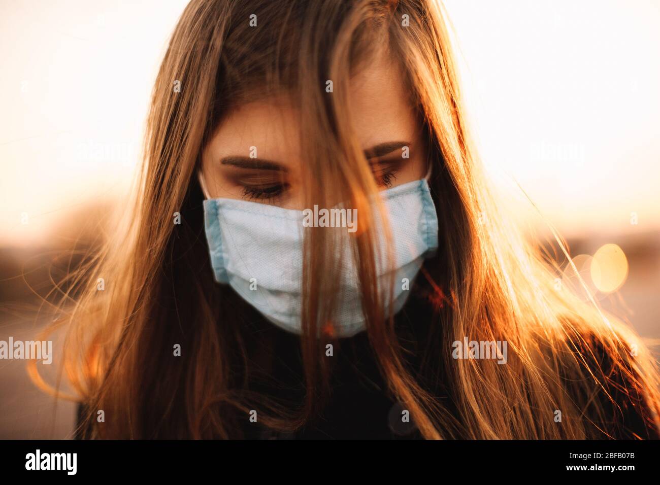Gros plan portrait de triste jeune femme déprimée portant un masque médical protecteur du visage en se tenant en ville pendant le coucher du soleil Banque D'Images