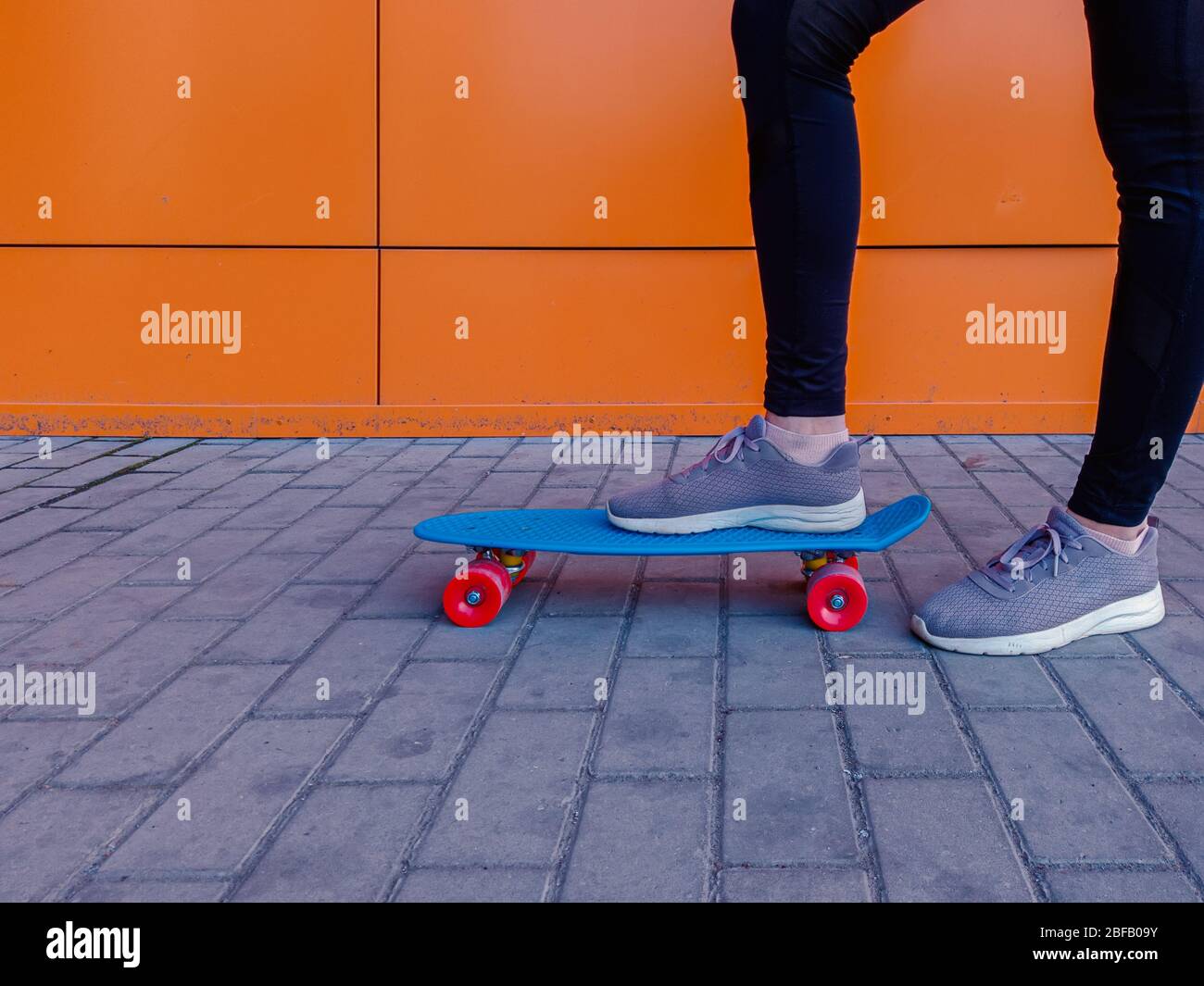 Photo d'une fille qui a mis son pied dans des baskets sur un skateboard contre un mur orange brillant Banque D'Images