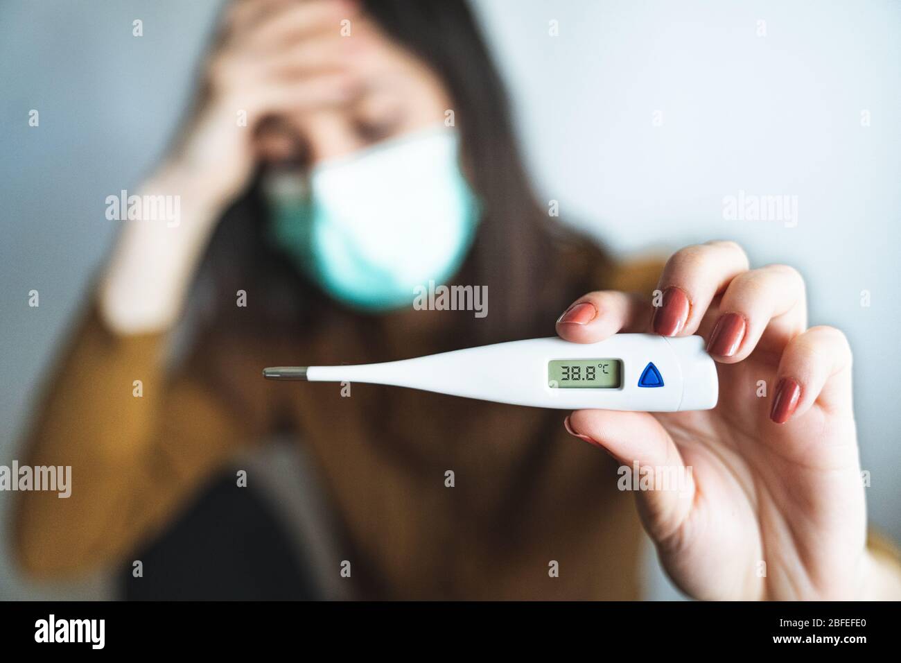 Foyer sélectif d'une jeune femme malade portant un masque chirurgical et tenant un thermomètre numérique qui indique qu'elle a plus de 38 degrés de fièvre. Malade et Banque D'Images