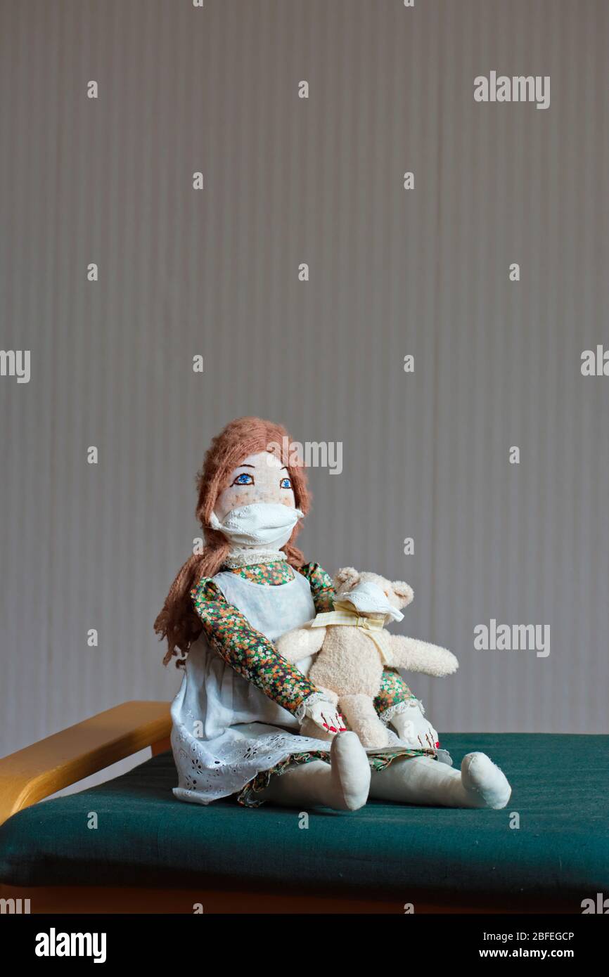 Une poupée féminine à l'ancienne faite à la main revêtue d'une robe agrémentée d'un robe chasuble est assise sur une chaise, portant un masque de protection. Elle tient un petit ours en peluche Banque D'Images