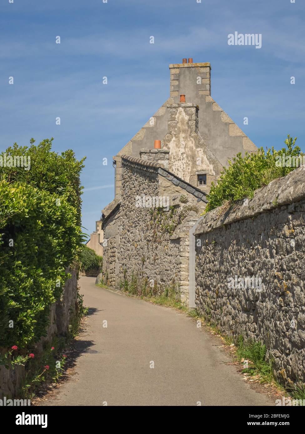 Les murs en pierre bordent une rue étroite et vide menant à un village avec de imposantes cheminées en Bretagne, en France, lors d'une journée d'été ensoleillée. Banque D'Images