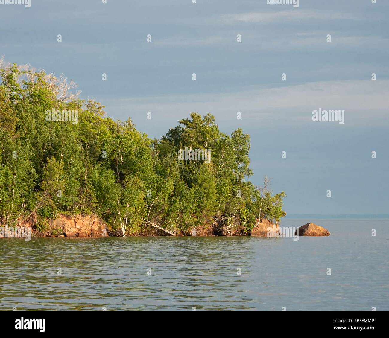 Point rocheux isolé avec lumière du soleil de la fin de l'après-midi sur une forêt entourée d'eau et de ciel bleu nuageux dans le Lakeshore national des îles Apôtre. Banque D'Images