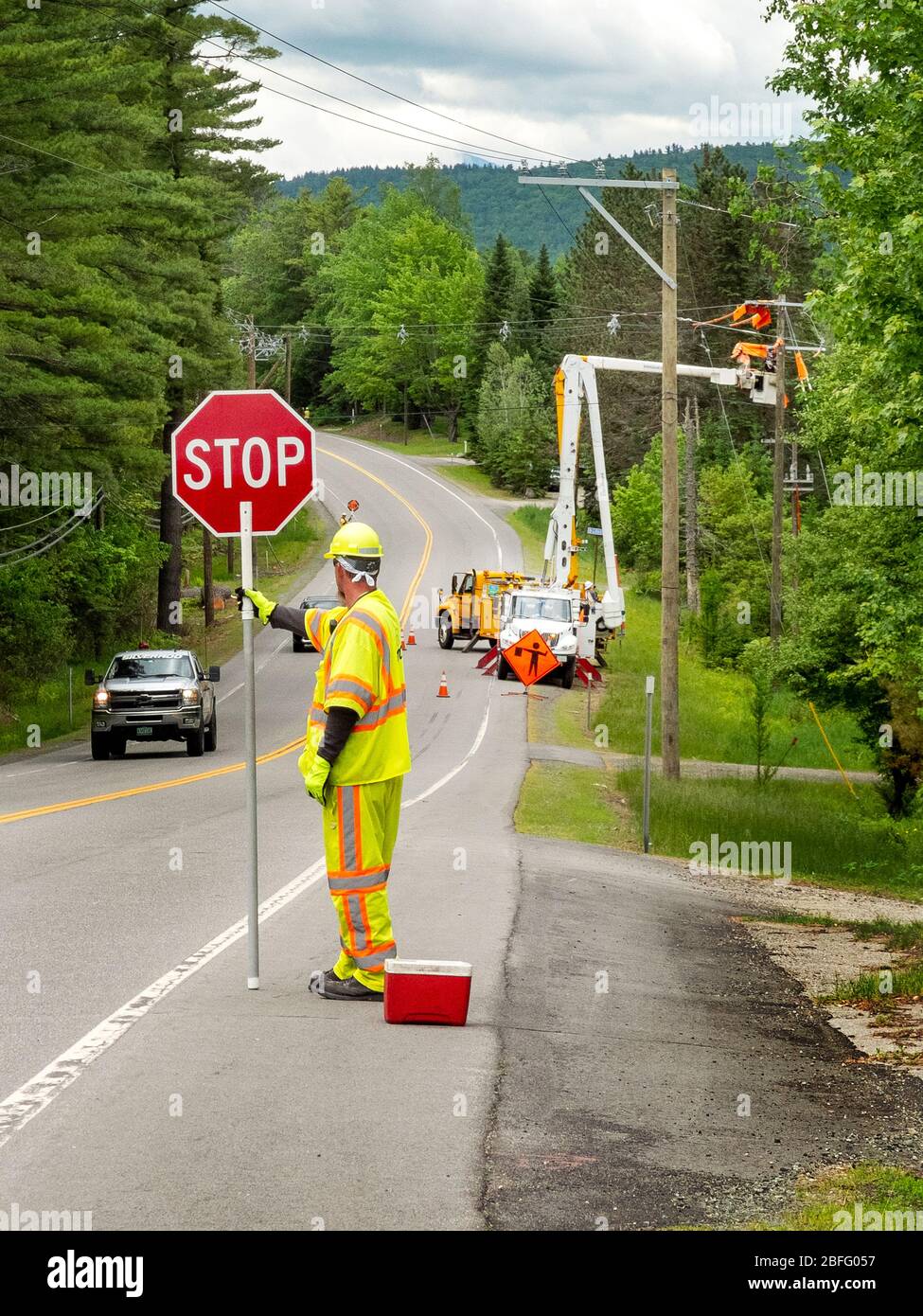 Un employé de l'état de la route possède un panneau d'arrêt pour contrôler la circulation sur un site d'emploi d'électriciens sur une grue de picker de cerisier qui répare les lignes de transmission électriques à Whitefield, dans le New Hampshire. Banque D'Images