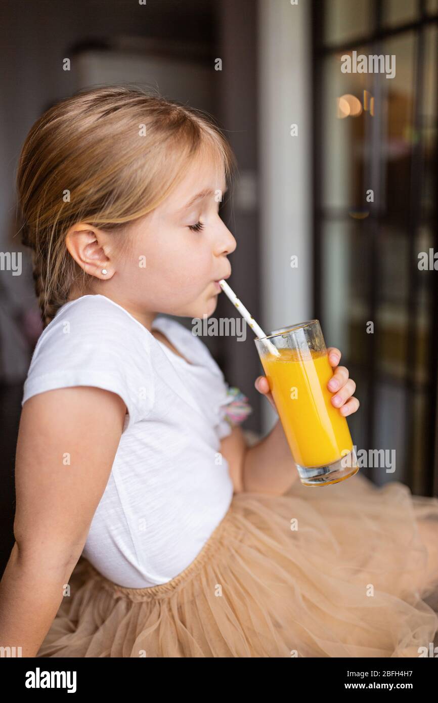 Jolie petite fille caucasienne avec cheveux blond assis sur la table de cuisine et boire du jus de mangue frais. Concept de mode de vie sain. Enfant de 6 ans Banque D'Images