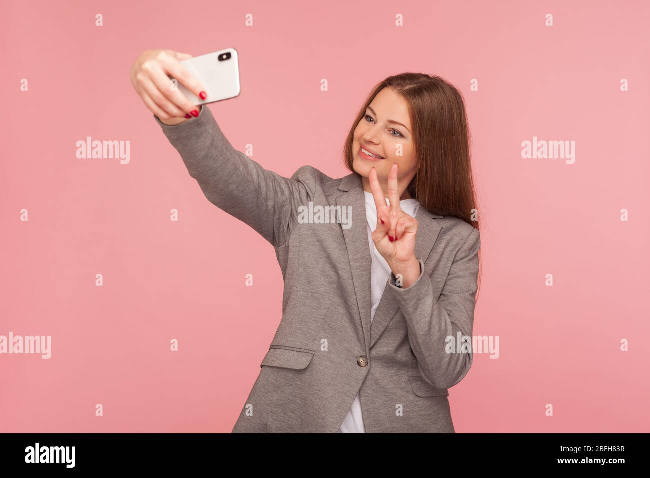 Portrait de femme d'affaires réussie dans la veste de costume passer un appel vidéo avec un geste de victoire, ayant une conférence en ligne à l'aide de téléphone portable, de cellulaire d'entreprise Banque D'Images