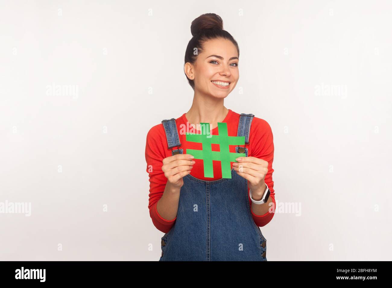 Suivez les tendances, Abonnez-vous à populaire blog! Portrait d'une jolie fille gaie et stylée avec un petit pain dans des salopettes en denim portant le symbole hashtag des réseaux sociaux Banque D'Images