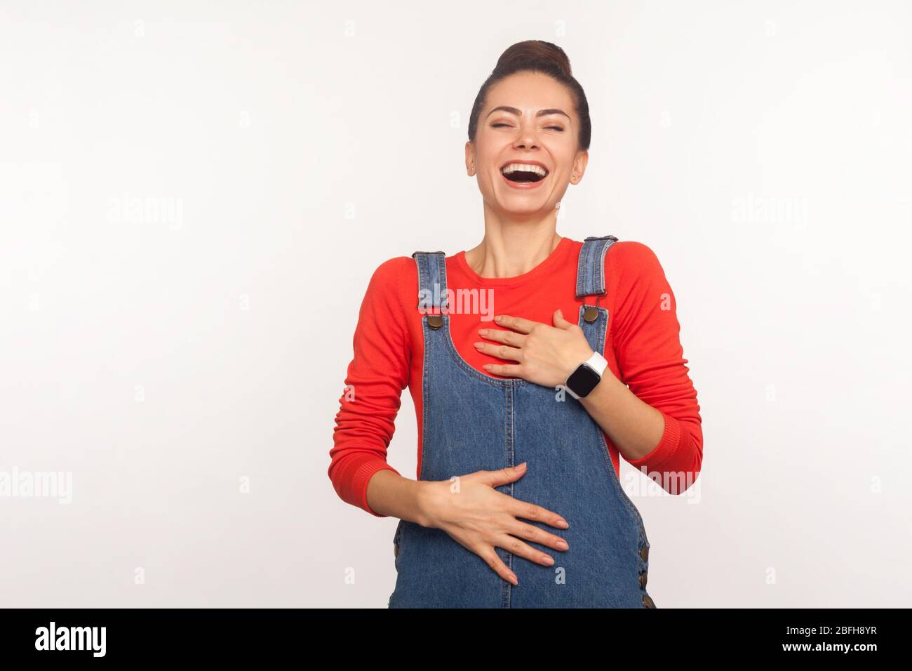 Rire hystérique. Portrait de positive heureuse fille élégante avec un pain de cheveux dans des salopettes en denim riant à fort et tenant le ventre, utilisé par le hum Banque D'Images