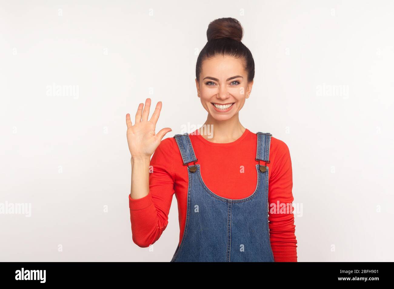 Portrait d'une jolie fille sympa et élégante avec un pain aux cheveux dans des salopettes en denim qui esturent un bonjour doux, accueillant avec un sourire toothy, se réveillant la main et saluant. Banque D'Images