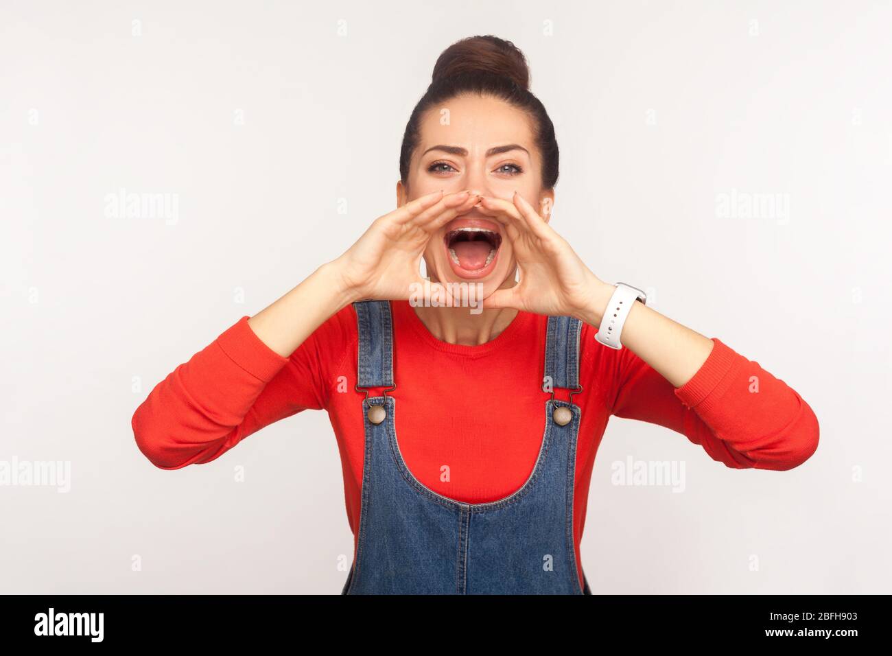 Attention, annonce! Portrait d'une fille élégante avec un pain à cheveux dans des combinaisons en denim tenant les bras près de la large bouche ouverte et message criant, annonce forte Banque D'Images