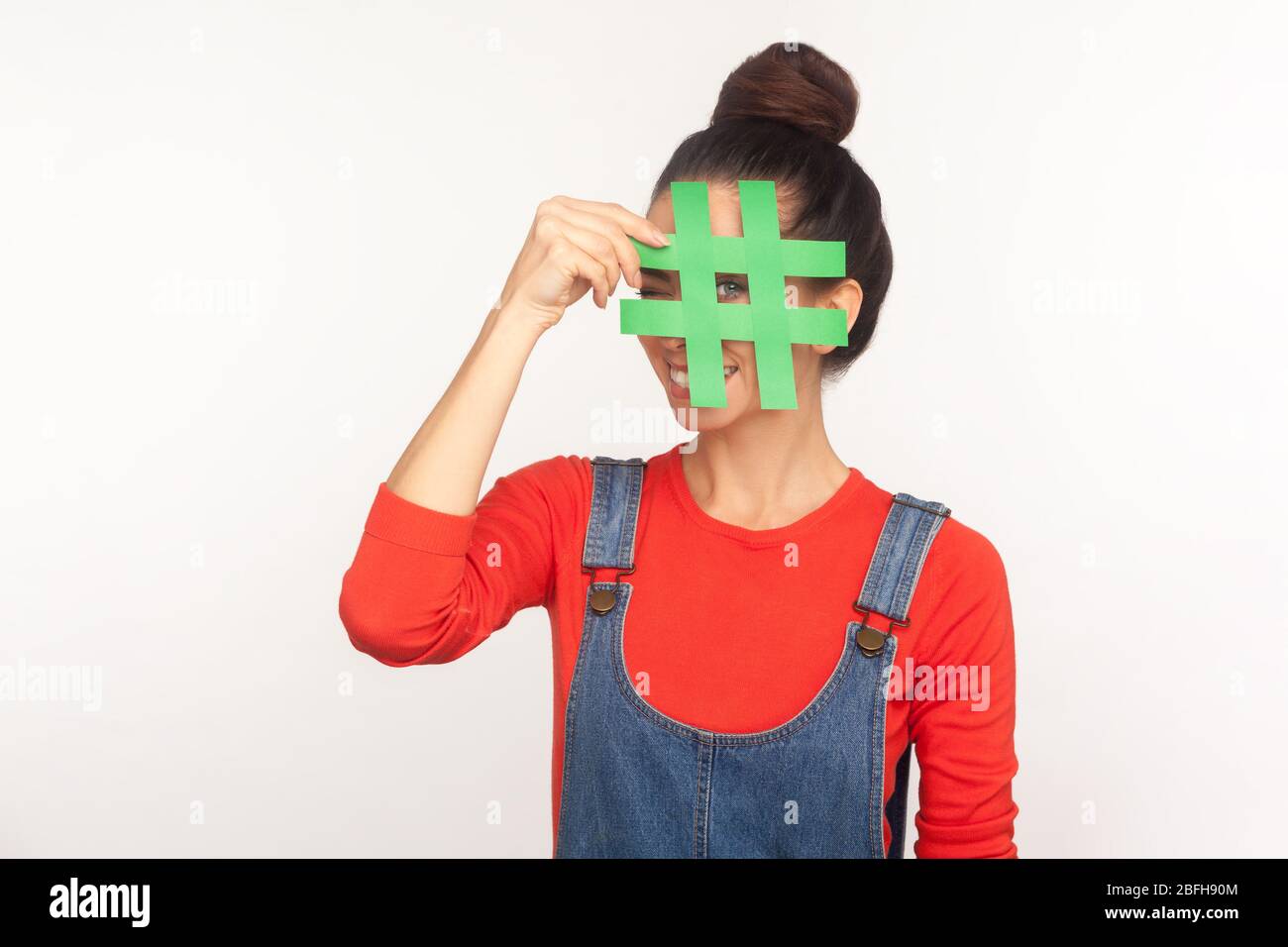 Portrait d'une jeune fille gaie avec un pain aux cheveux dans des combinaisons en denim couvrant le visage avec le symbole hashtag des médias sociaux, recommandant de suivre le contenu à la mode, popul Banque D'Images